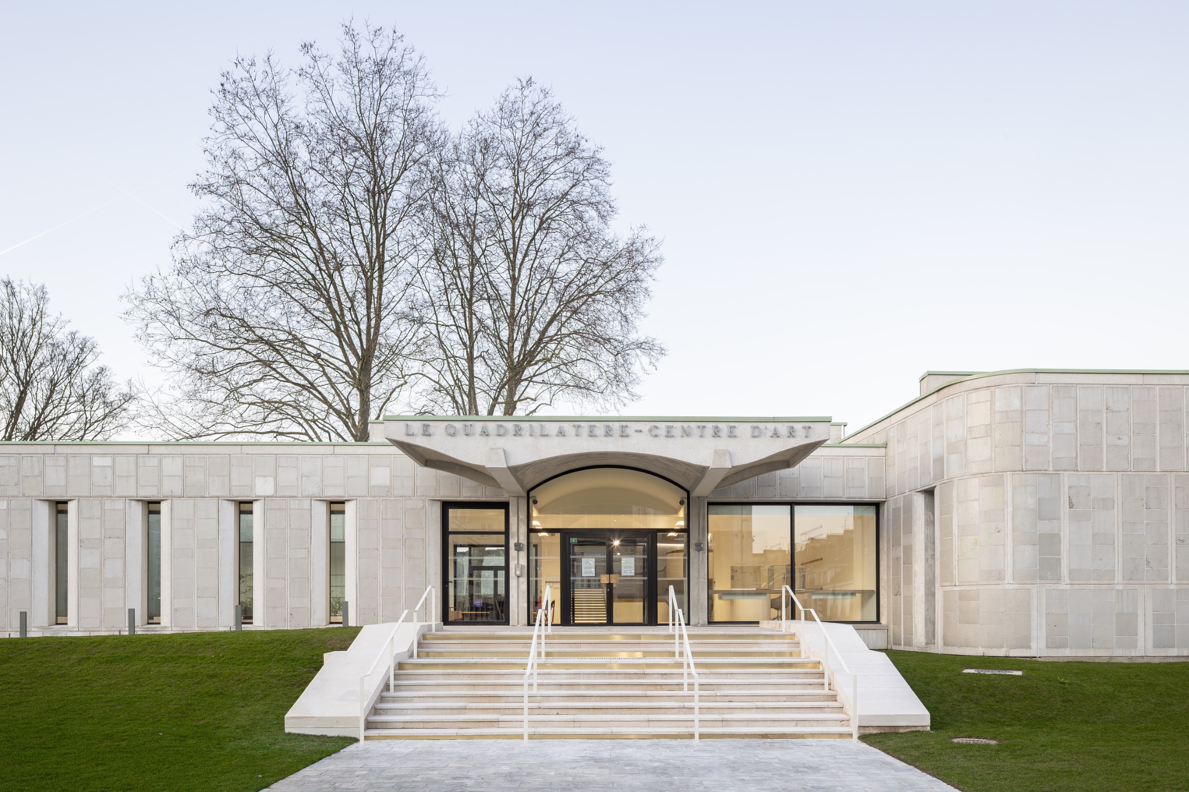 Bâtiment moderne en pierre avec entrée en escalier et large vitrine. Arbre à l'arrière.